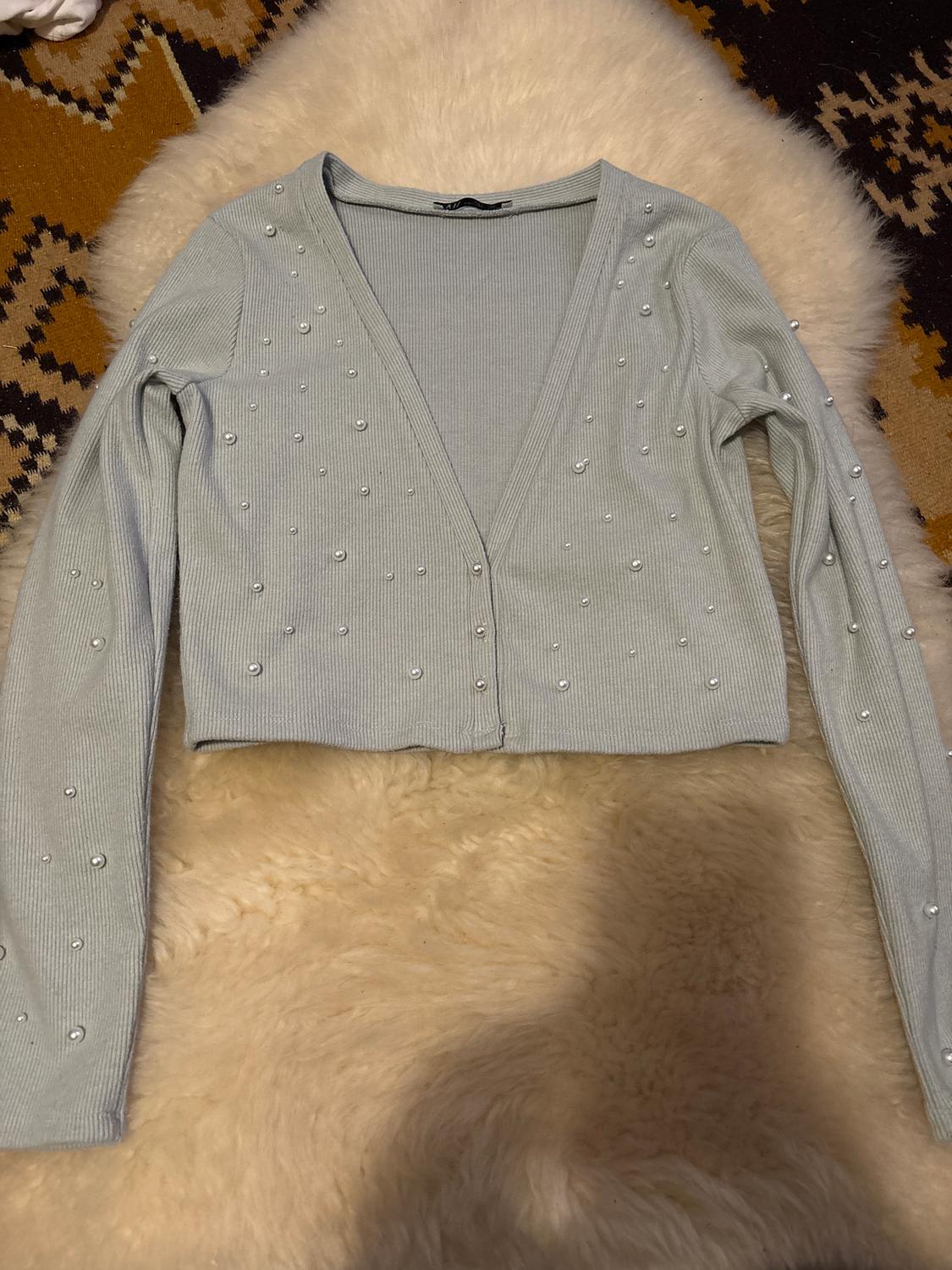 Light grey cardigan with decorative pearls on a geometric pattern rug and white faux fur background.