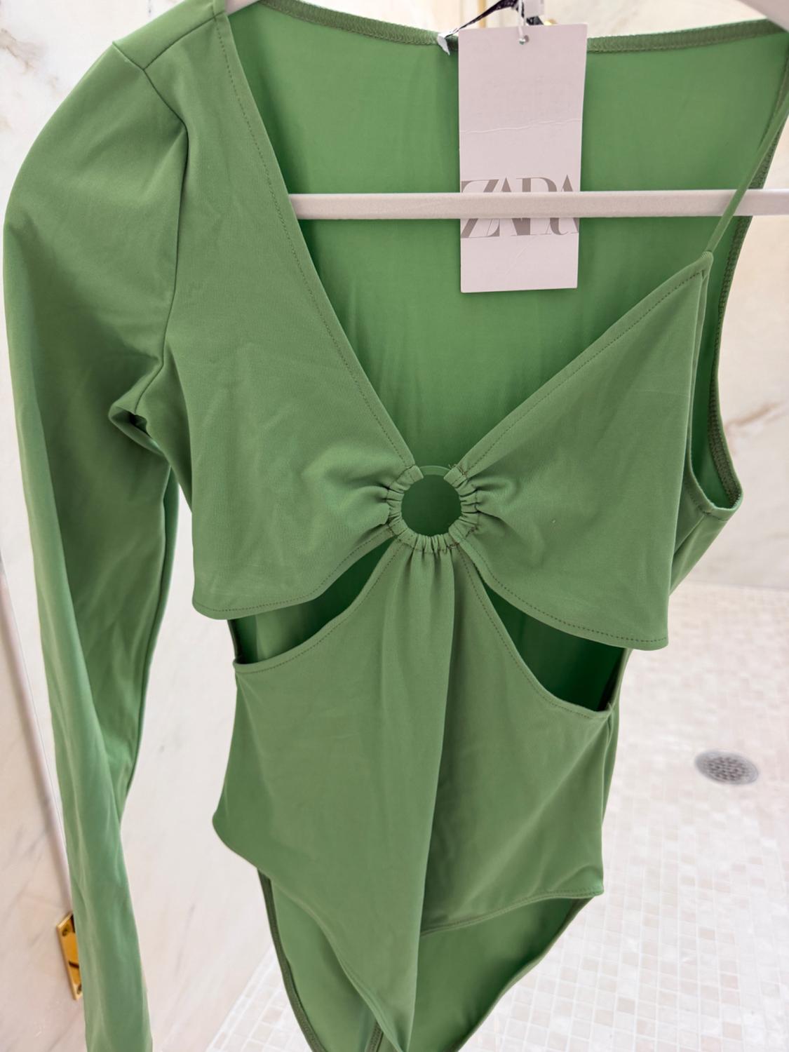 Green bodysuit with cut-out design and central ring, hanging on a hanger. Visible label with "ZARA" text. Light tiled background.