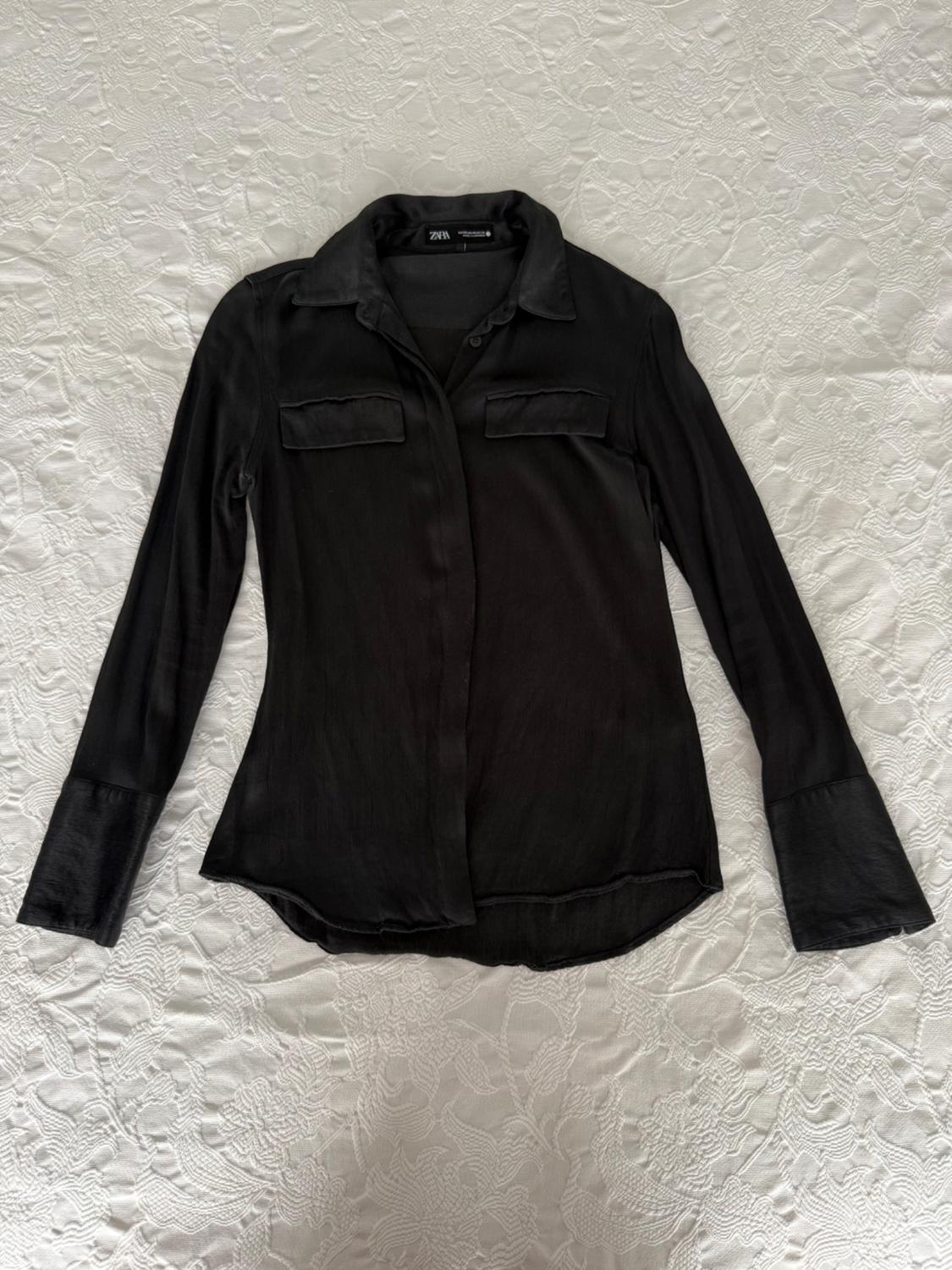 Black long-sleeved shirt with front pockets, spread out on a textured white fabric. Label with ZARA text.
