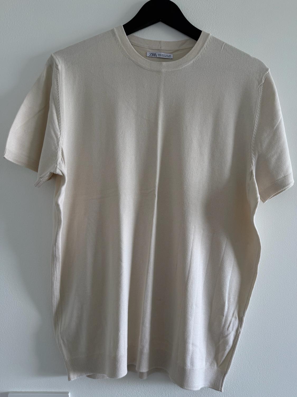 Beige knit short-sleeve t-shirt hanging on a black hanger against a white wall. Tag with the text zara.