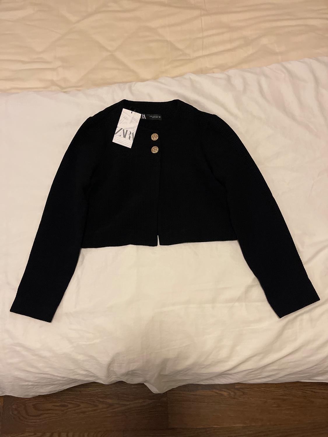 Black long-sleeved jacket with two gold buttons and a visible label on a bed with white and beige sheets.