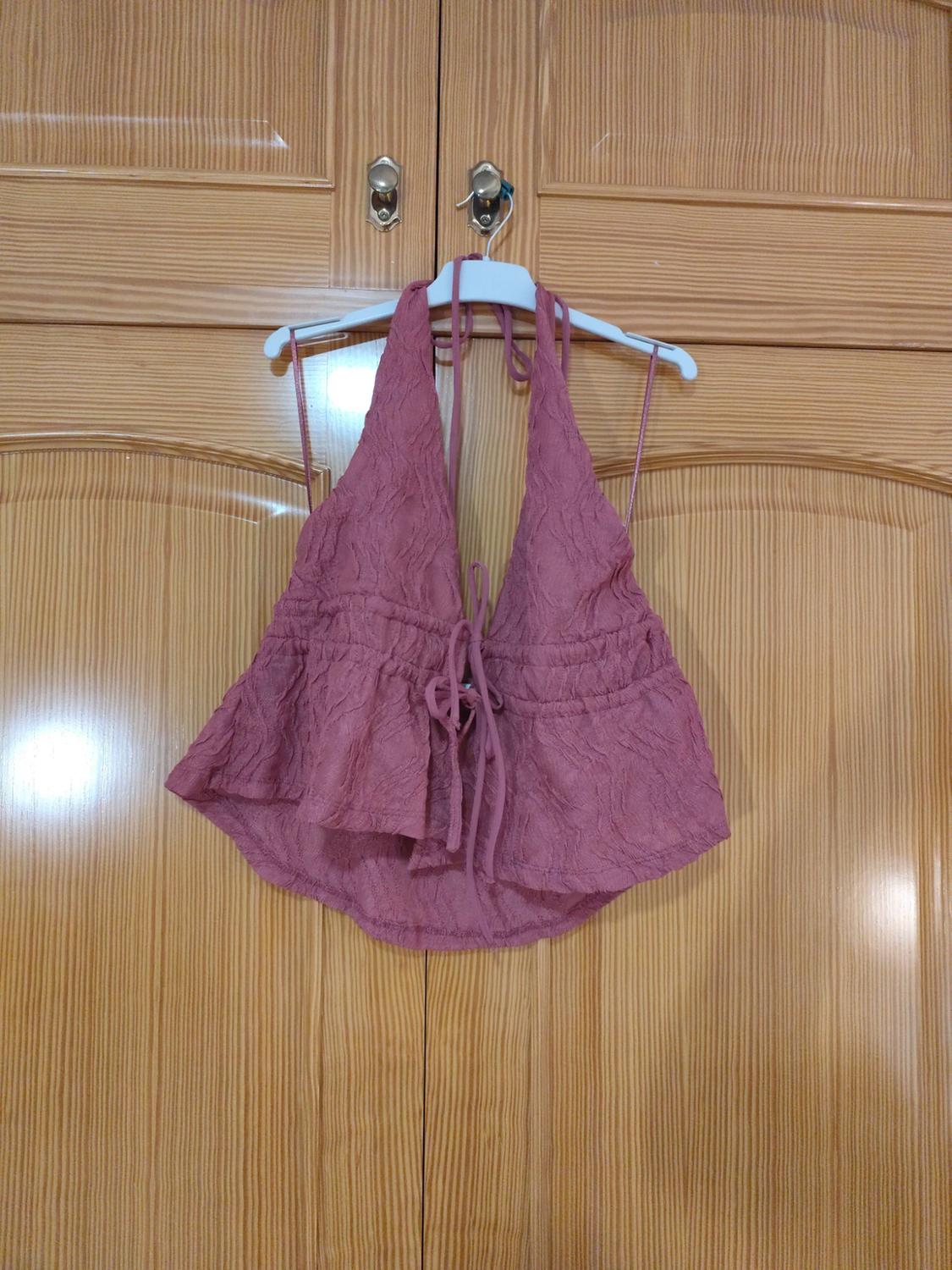 Casual ροζ τοπ με halter λαιμόκοψη, ανάγλυφο ύφασμα και φιόγκο μπροστά, κρεμασμένο σε λευκή κρεμάστρα πάνω από ξύλινη ντουλάπα.