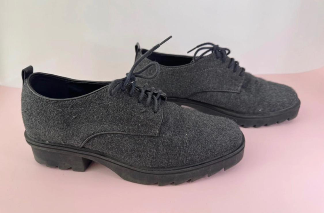 Dark grey felt shoes with black laces and a thick black sole, on a light pink and white background.