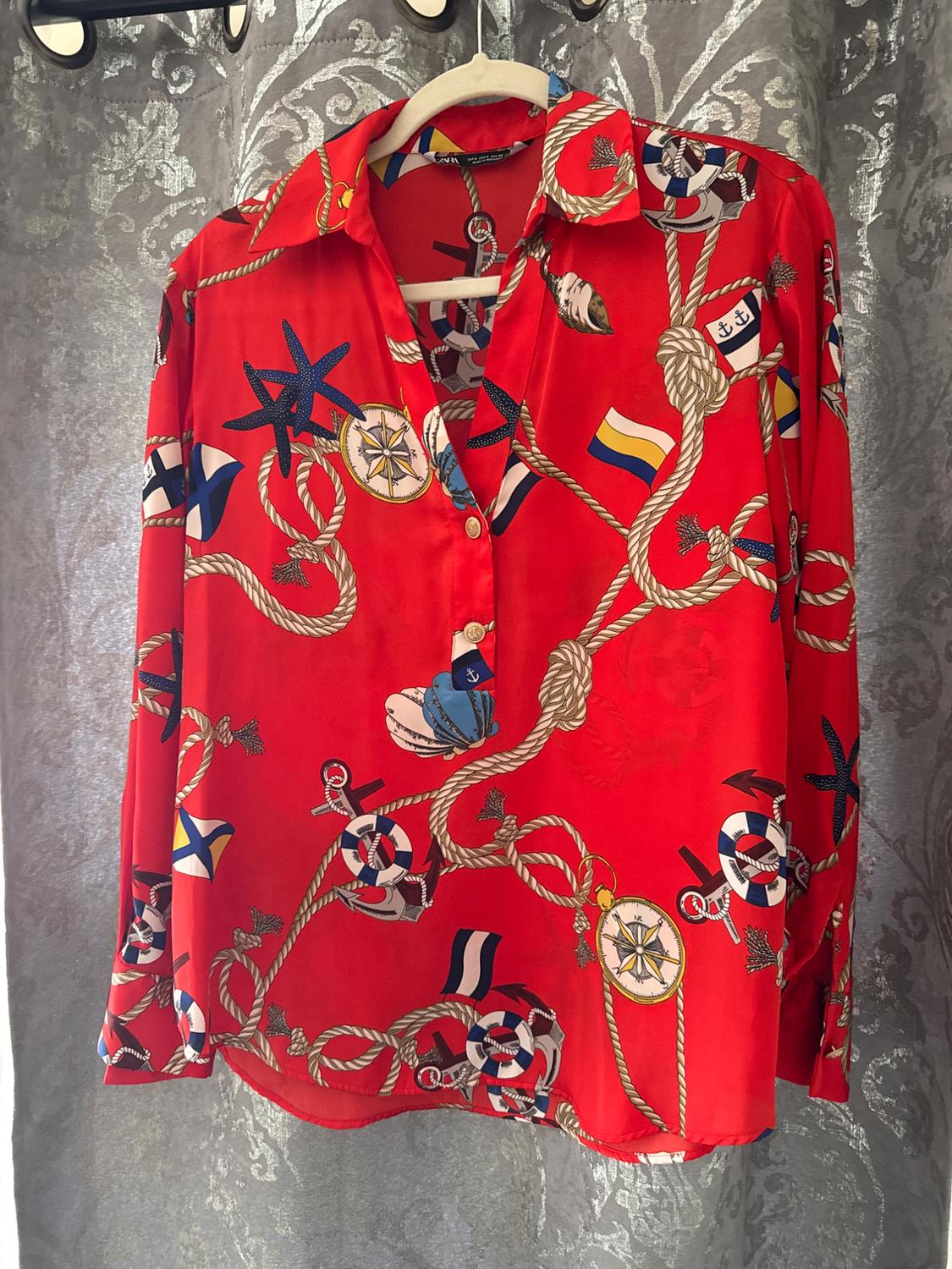 Red shirt with a nautical print of ropes, compasses, and life preservers, hanging on a hanger against a gray curtain background.