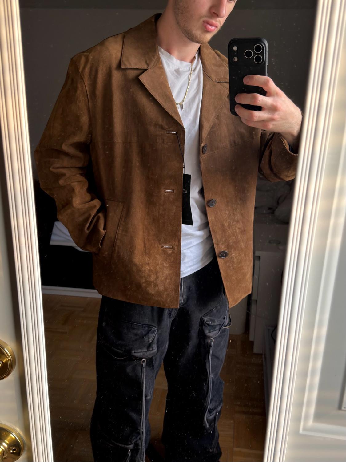 Man taking a selfie in a mirror. He is wearing a brown jacket, white T-shirt, necklace, and black cargo pants with pockets. Indoors.