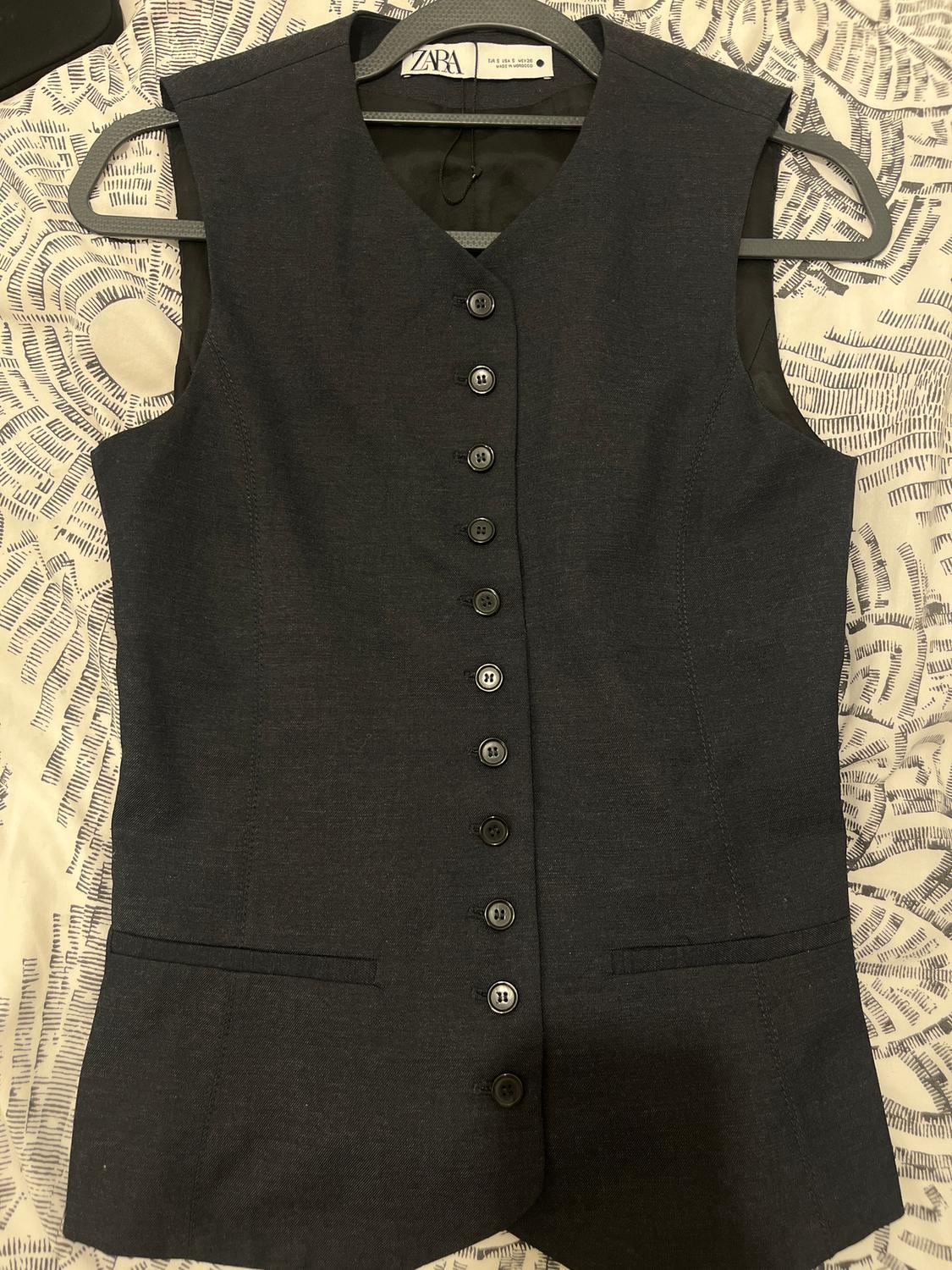 Black sleeveless waistcoat with front buttons, hanging on a hanger over a patterned fabric. Visible label: Zara.