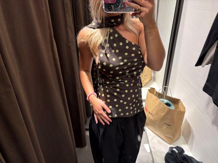 Selfie in a changing room. Woman wearing a dark, asymmetric polka-dot top, a neck scarf and black trousers. A paper bag can be seen in the background.