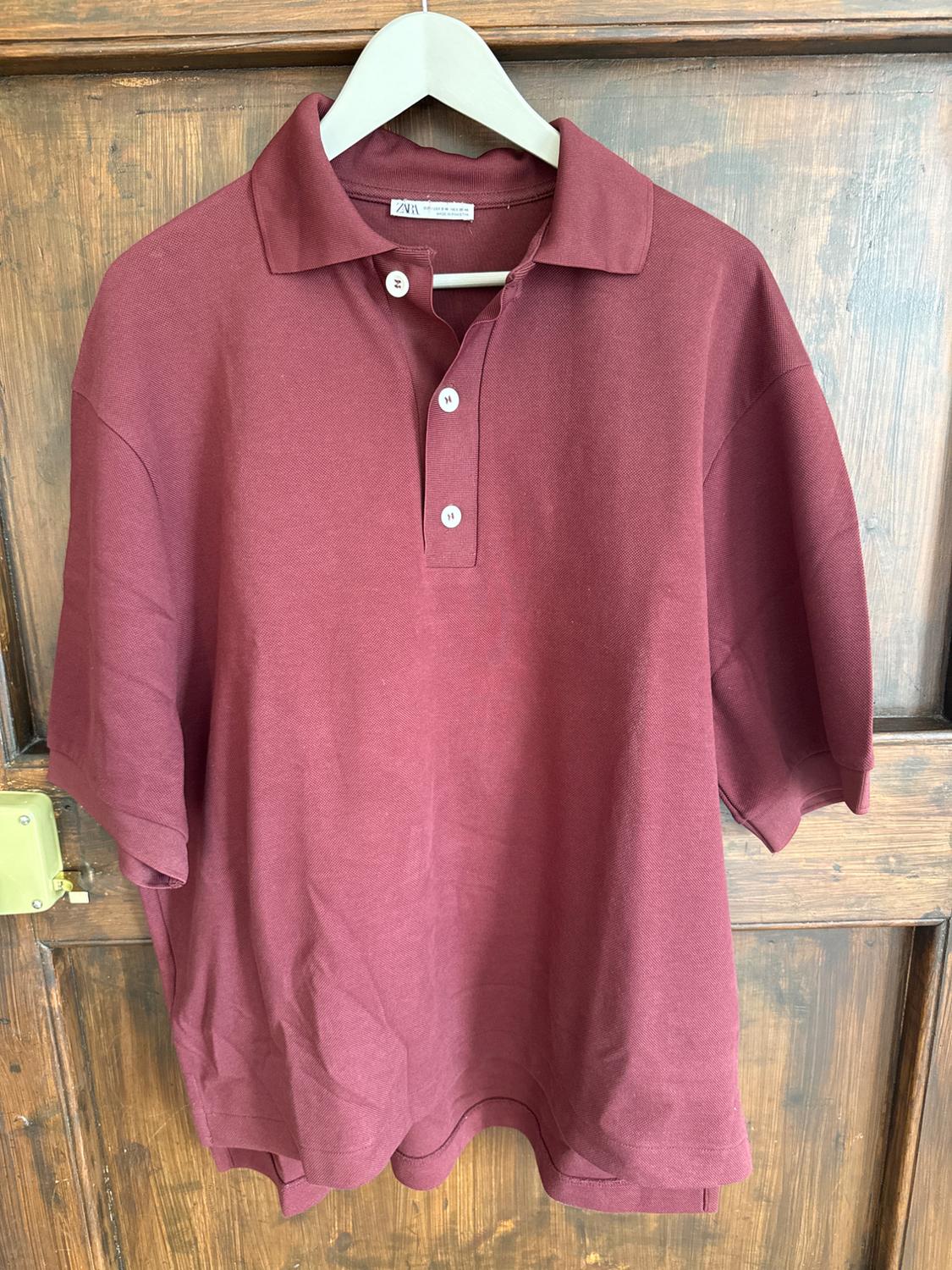 Burgundy short-sleeved polo shirt with three white buttons, hanging on a white hanger over a wooden door.