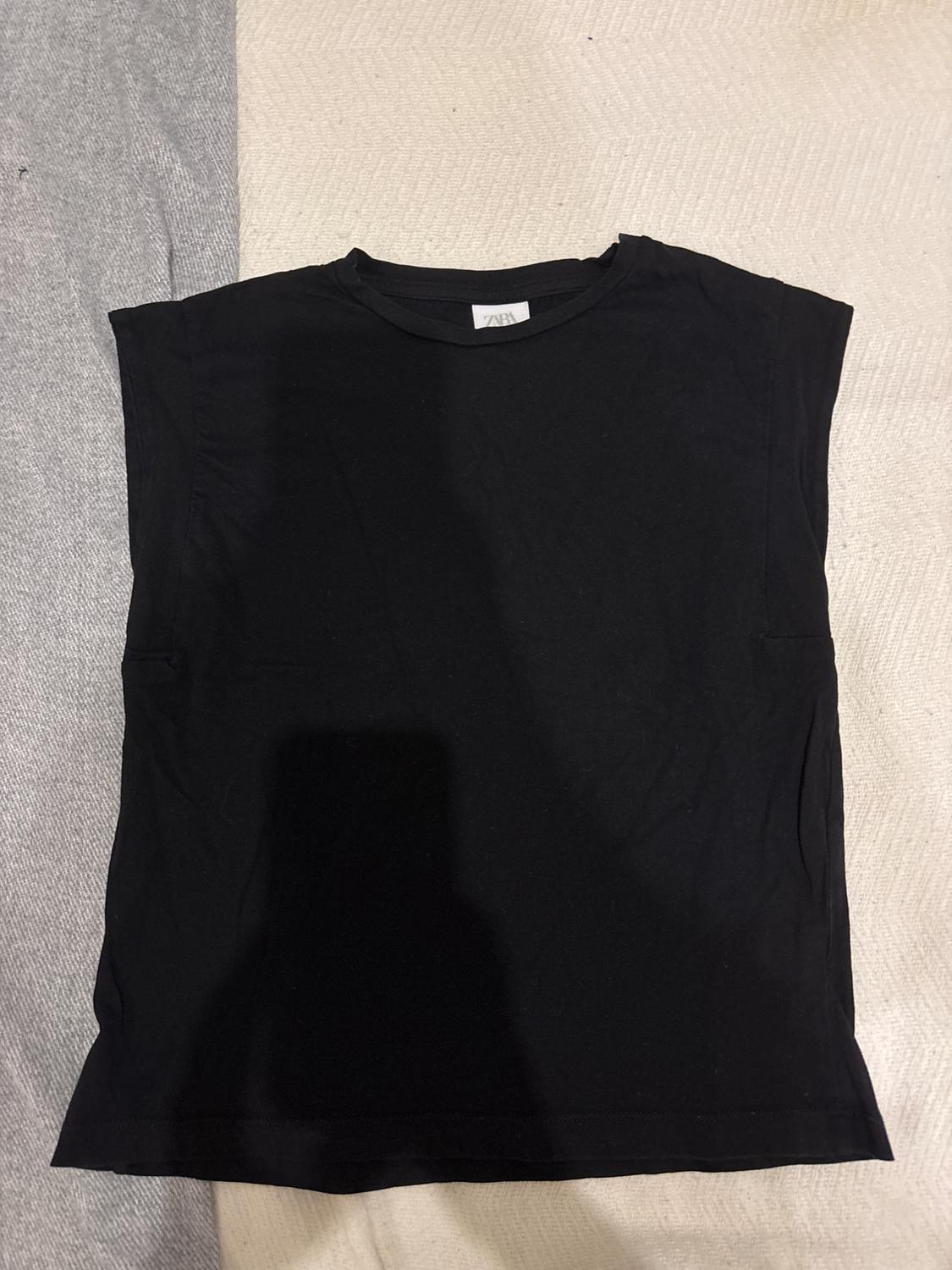 Sleeveless black t-shirt on a grey and beige fabric surface. Label visible on the neck with text "ZARA".