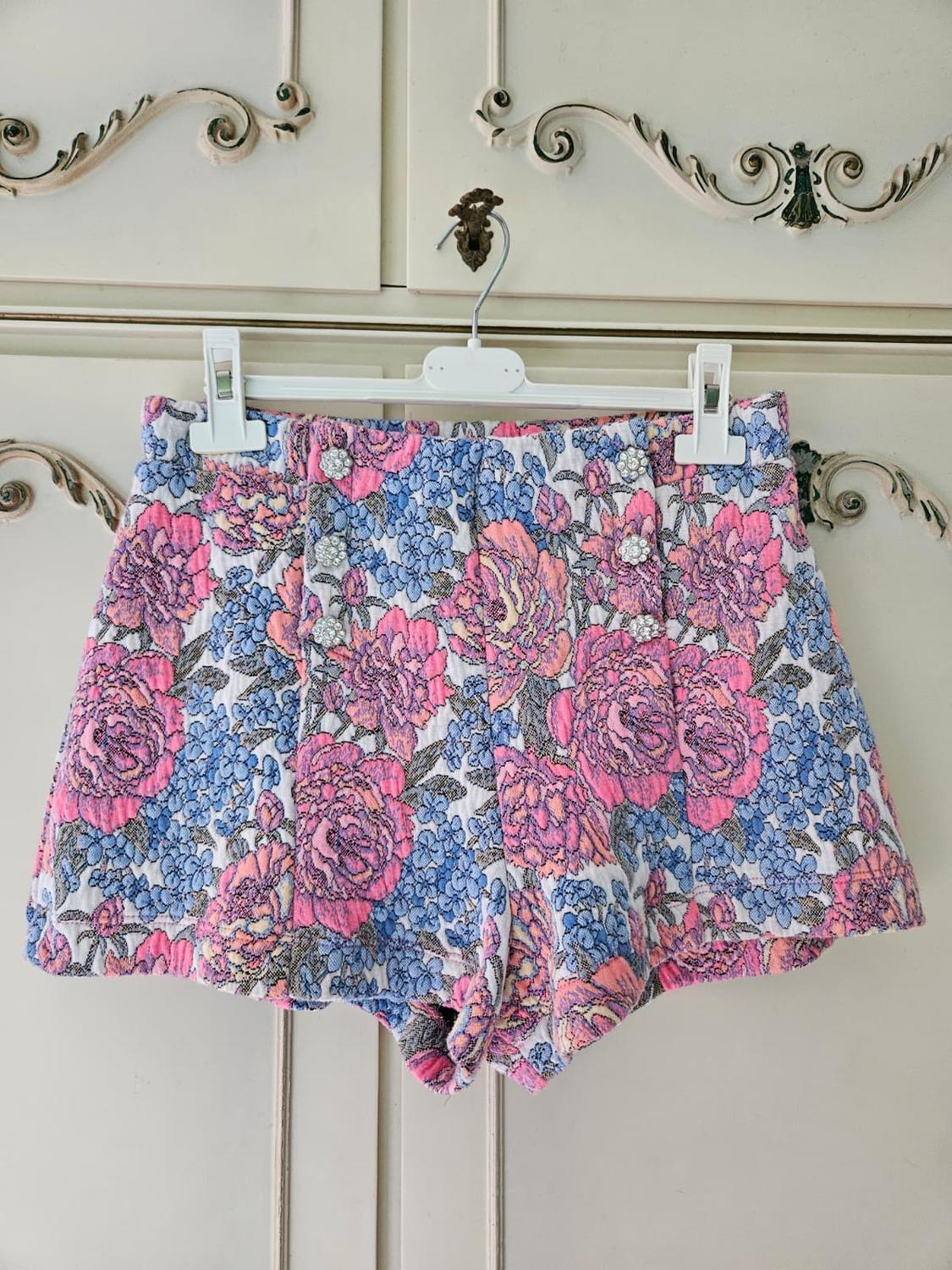 High-waisted shorts with a pink and blue floral print and jewelled buttons, hanging on a hanger in front of a vintage-style white wardrobe.