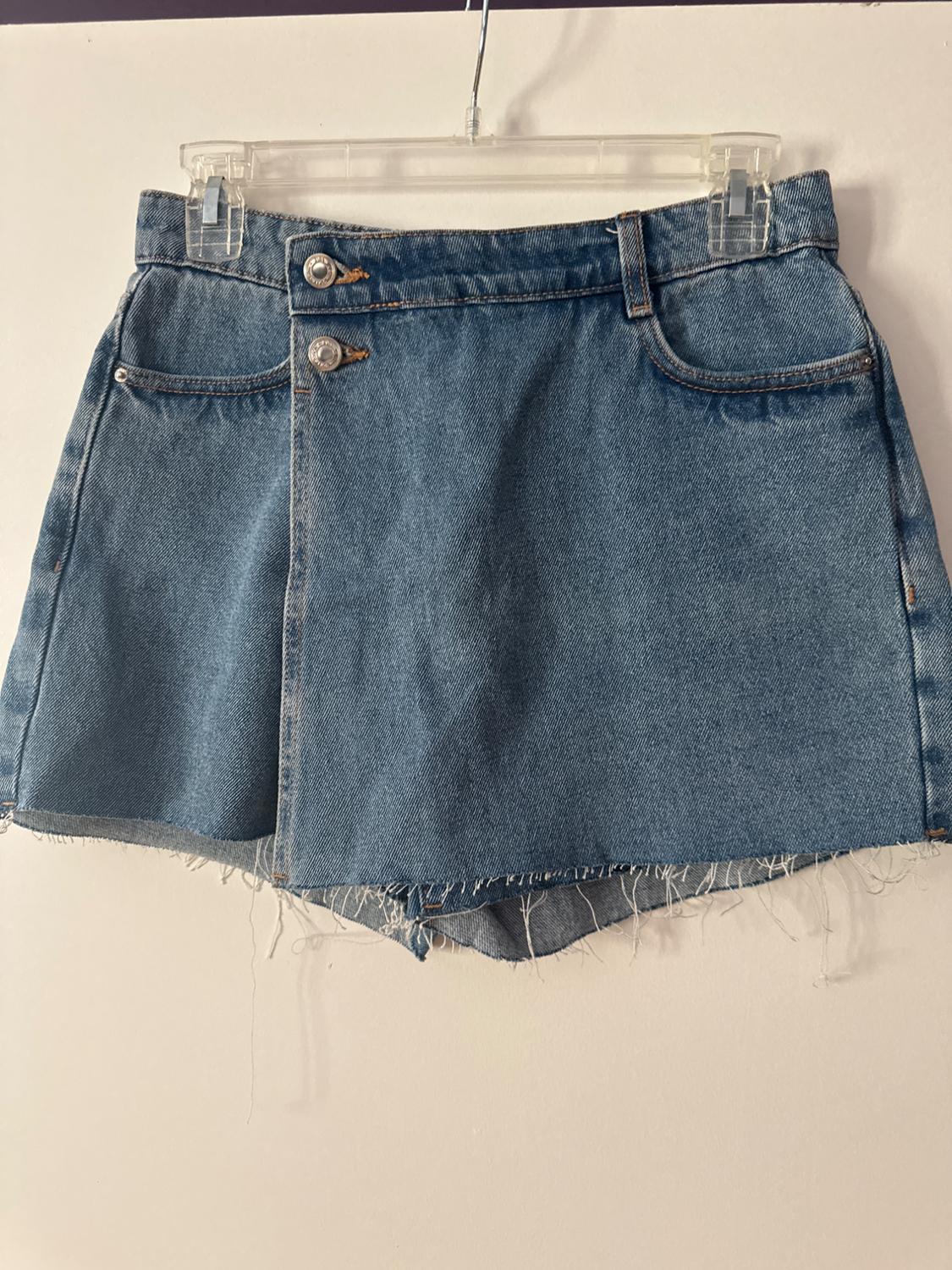 Blue denim wrap skort with buttons and a frayed hem, hanging on a clear hanger against a white wall.