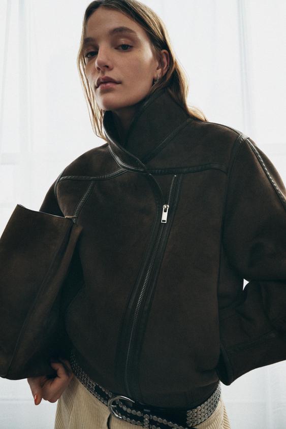 Střední detail tmavě hnědé bundy se stojáčkem, předním zipem a dlouhými rukávy. Skryté kapsy ve švu.