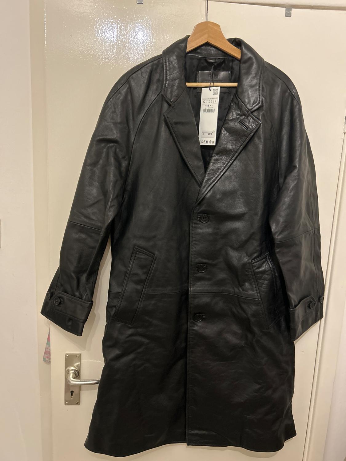 Black leather coat hanging on a wooden hanger, with visible tag. White door background with metallic knob.