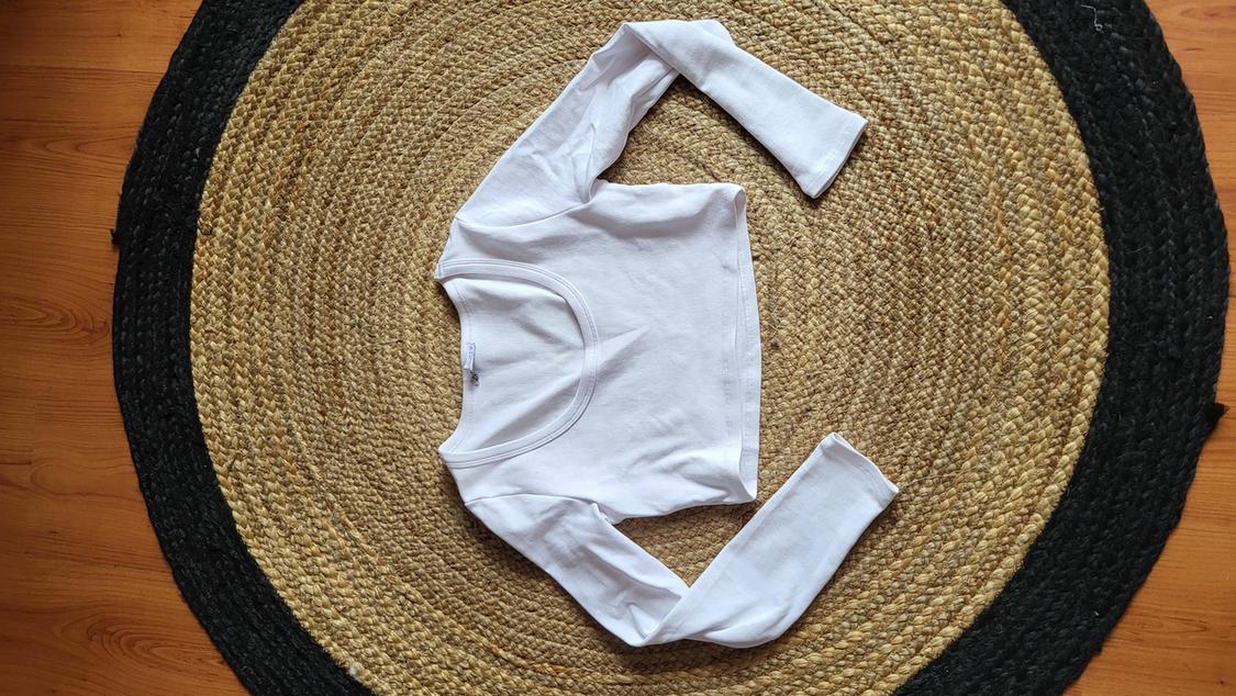 White long-sleeved T-shirt on a circular black and beige jute rug, placed on a wooden floor.