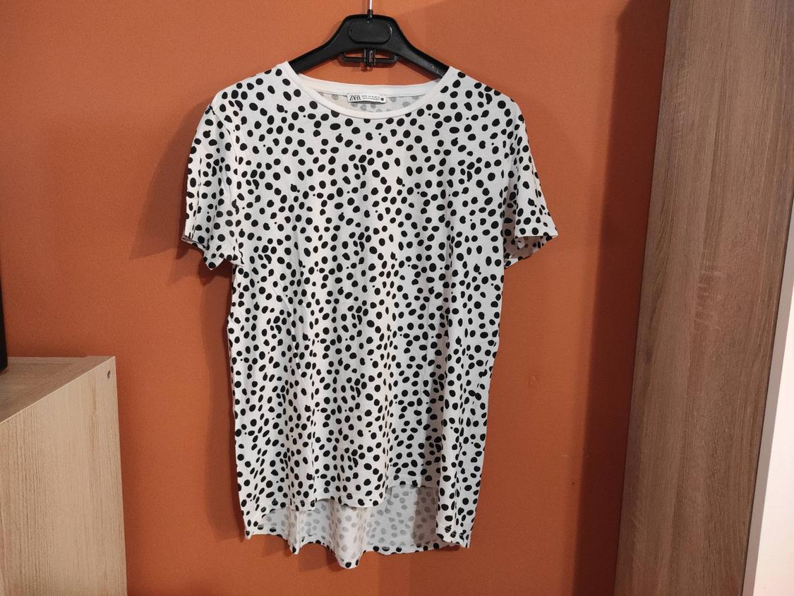 White t-shirt with black polka dot print hanging on a black hanger against an orange wall.