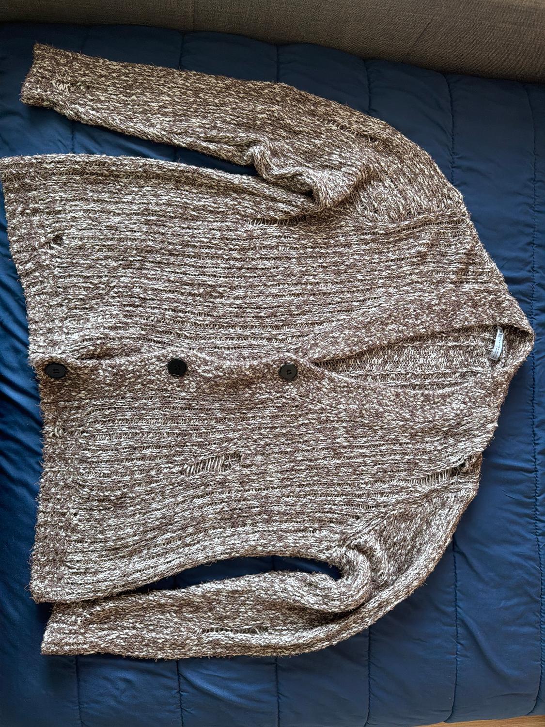 Brown knitted cardigan with black buttons on a blue quilted surface. Textured and relaxed design.