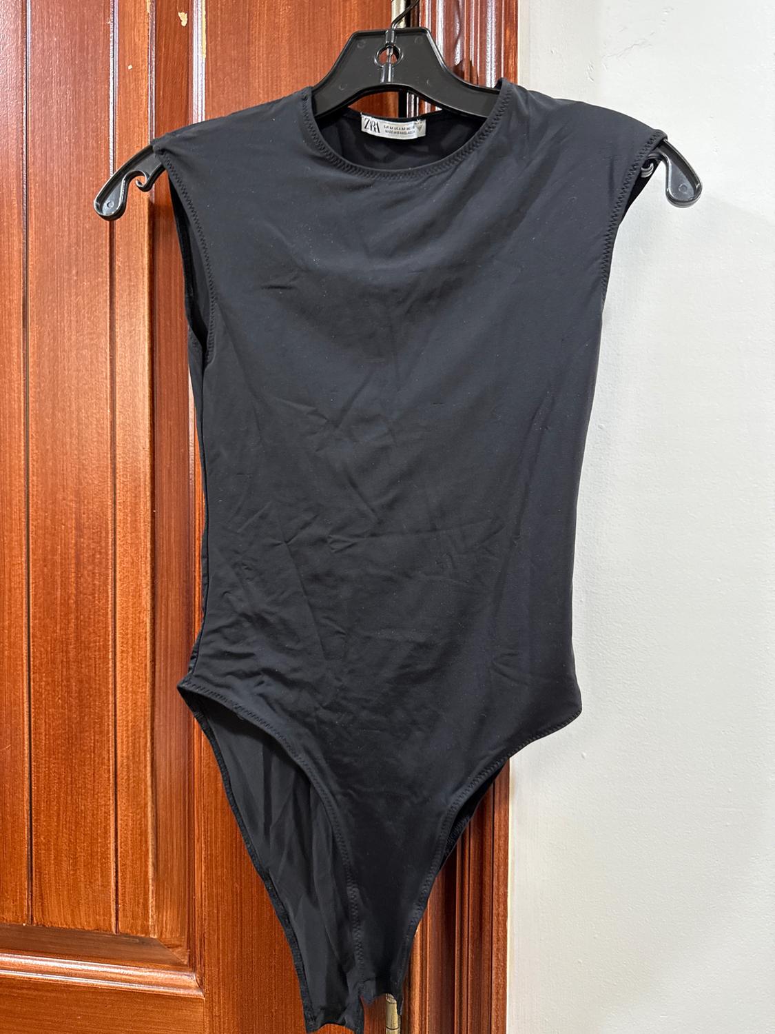 Sleeveless black bodysuit hanging on a black hanger on a wooden door. Label visible on the neck.