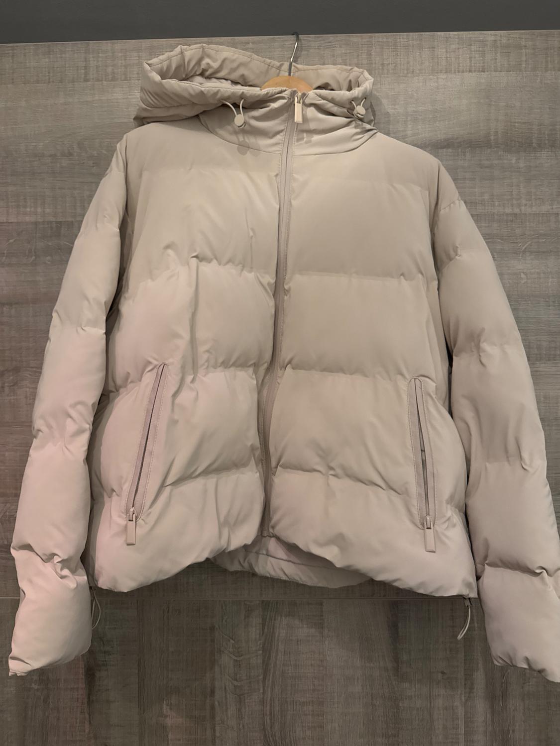 Beige padded jacket with hood and front zip, hanging on a hanger against a grey wooden background.
