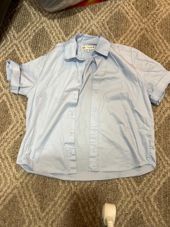 Light blue short-sleeved shirt with a classic collar, wrinkled, on a beige rug with a geometric pattern. Visible tag: ZARA.