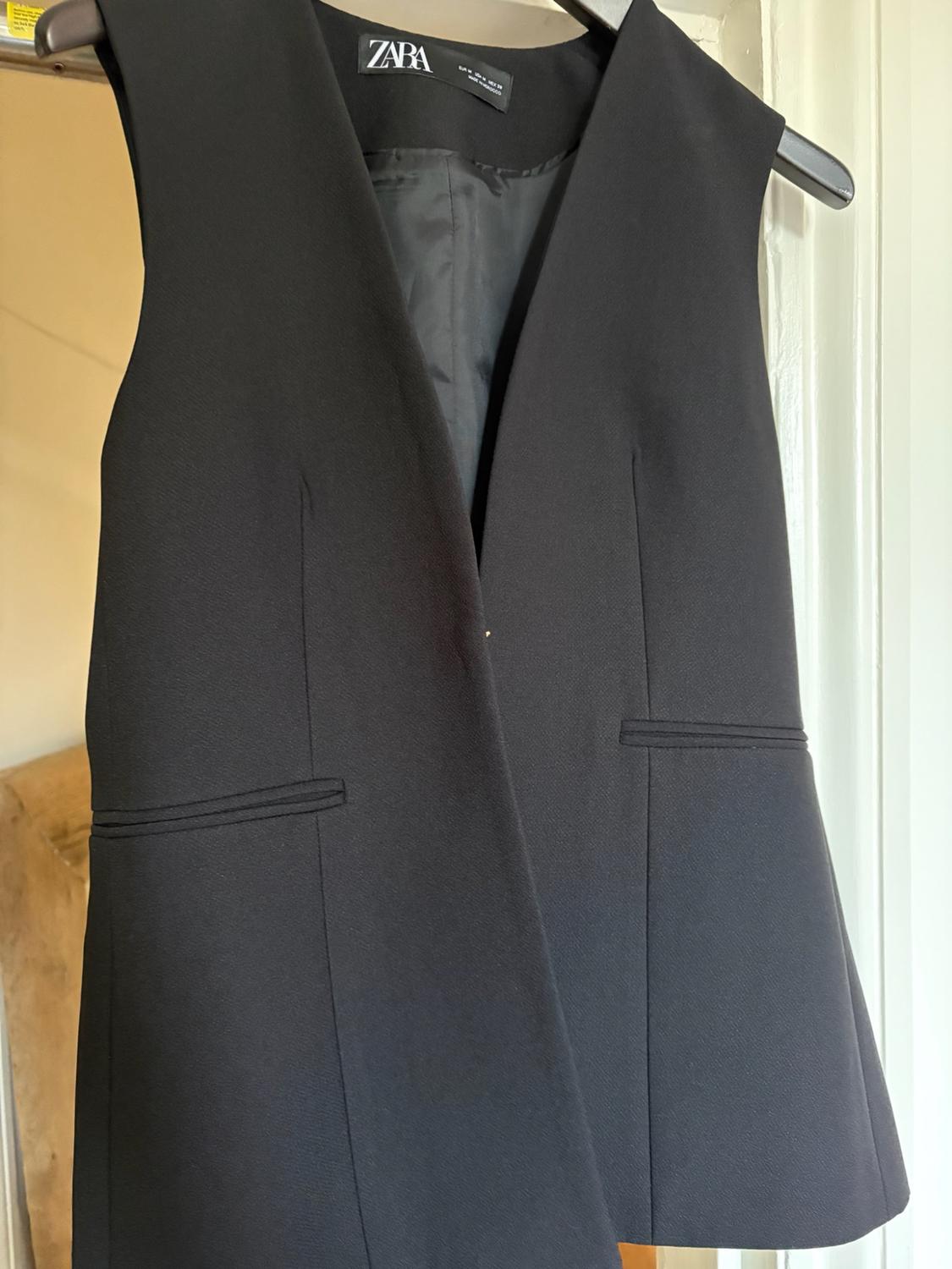 Black sleeveless waistcoat hanging on a hanger. V-neck design, front pockets and visible ZARA label on the inside.
