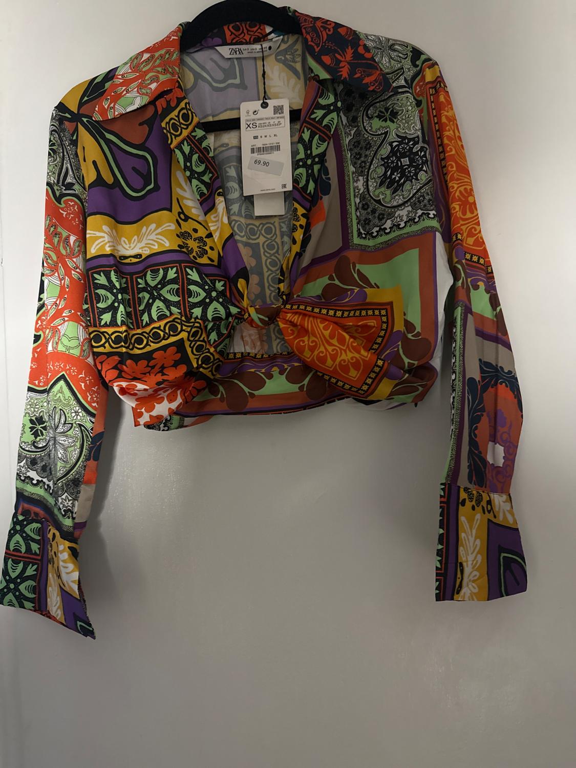 Colorful blouse with geometric and floral print, hanging on a hanger. Visible tag with price of 69.90 and size XS.