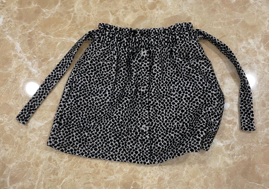 Short skirt with black and white speckled print, gathered elasticated waist, tie detail and front buttons on a beige marble background.