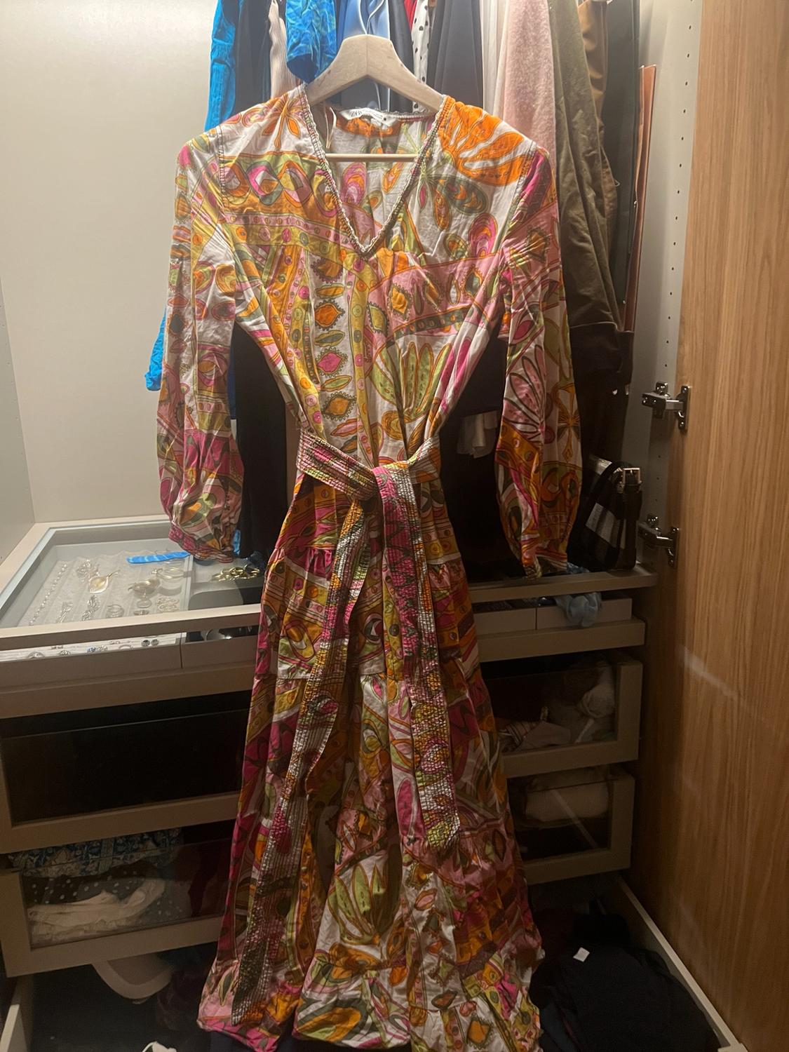 Long dress with a multicoloured print, V-neck and belt, hanging on a wooden hanger inside a wardrobe.