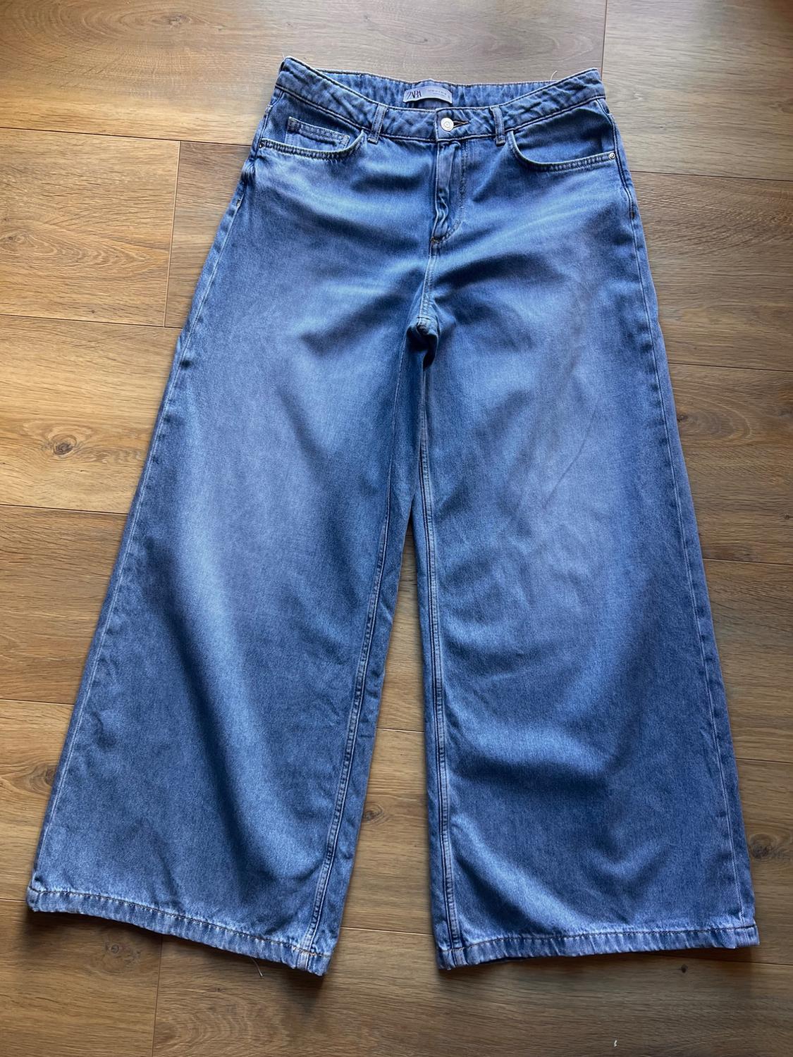 Blue denim wide-leg jeans on a light wooden floor. High-waisted design with front and back pockets.