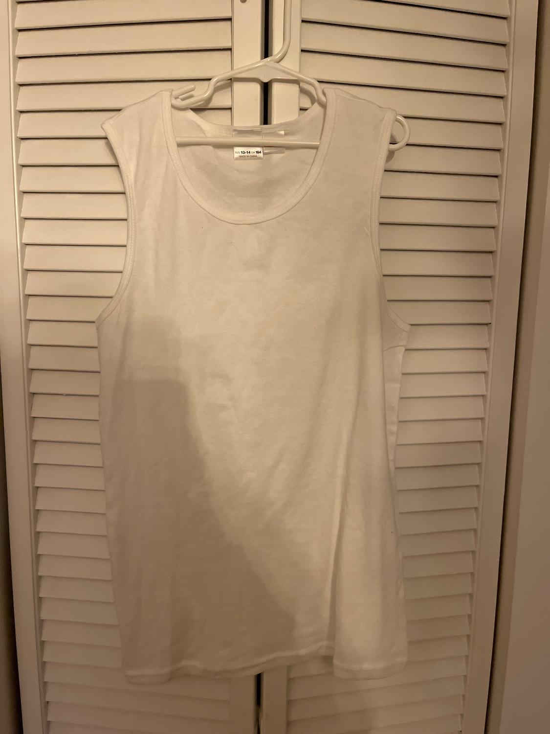 White sleeveless top hanging on a hanger in front of white louvered doors. Visible label with size 13-14 years.