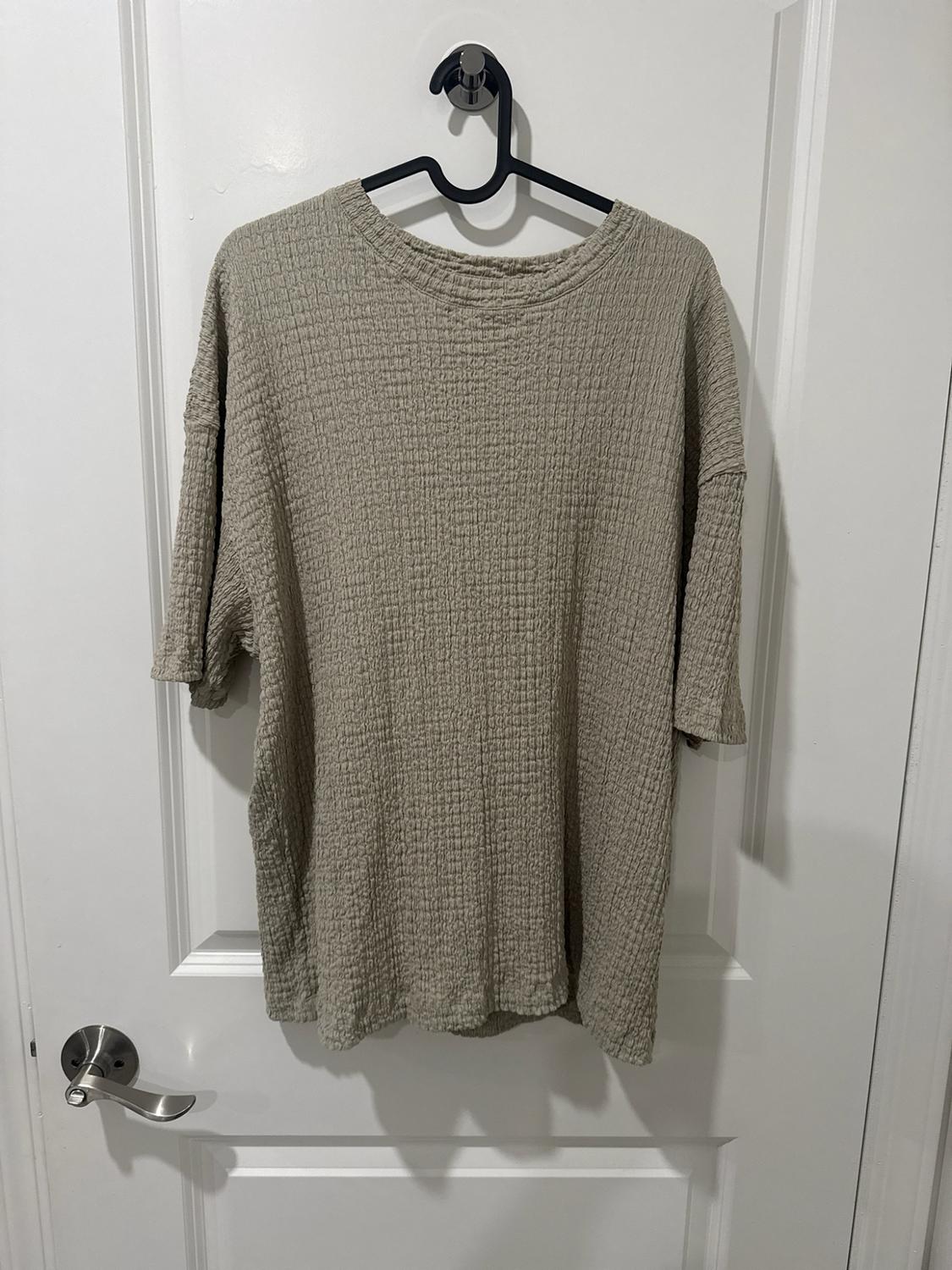 Loose beige short-sleeve T-shirt with a rough texture, hanging on a black hanger on a white door with a doorknob.