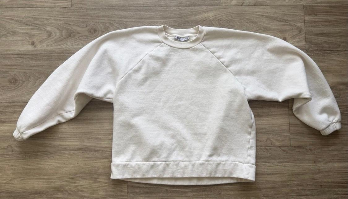 Cream-coloured plain crew-neck sweatshirt with wide sleeves, laid flat on a light wood floor. Relaxed style composition.