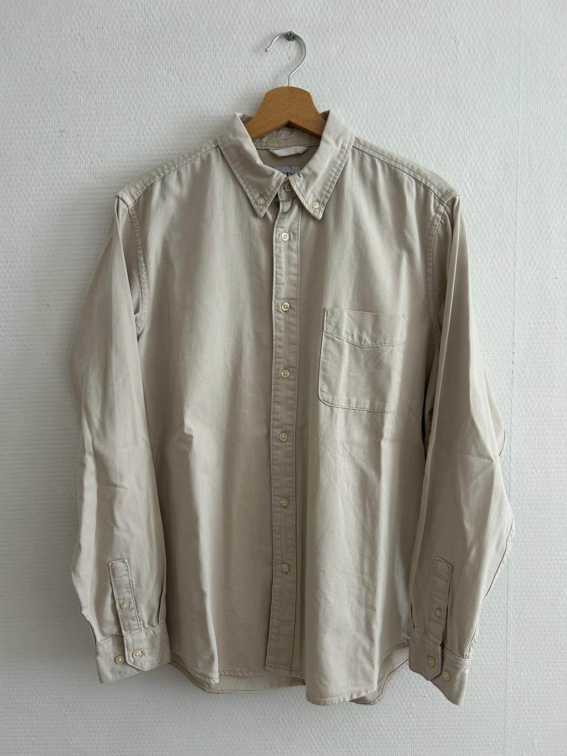 Beige long-sleeved shirt hanging on a wooden hanger against a white wall. It has a chest pocket and light-coloured buttons.