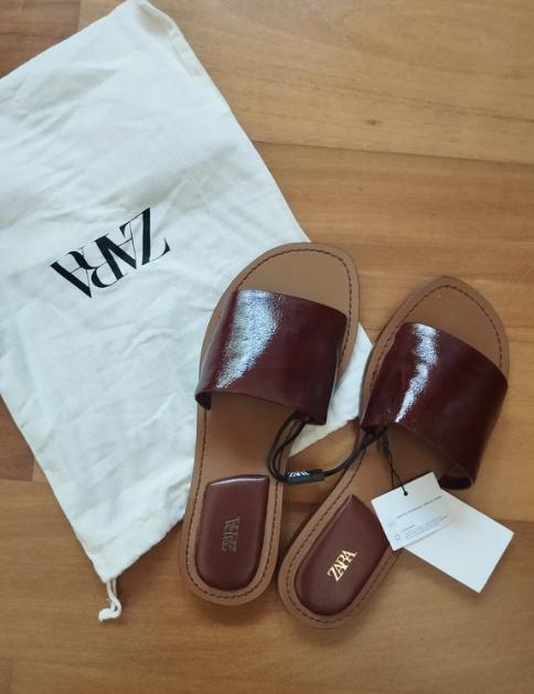 Brown wide-strap sandals on a wooden floor, next to a white bag with a black "ZARA" logo. Labels hanging.