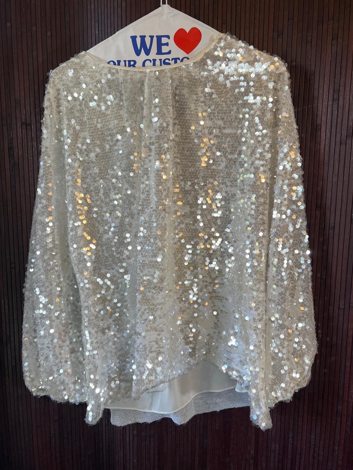Silver sequin cape hanging on a hanger with "WE ❤️ OUR CUSTOMERS" text on a dark wooden background.