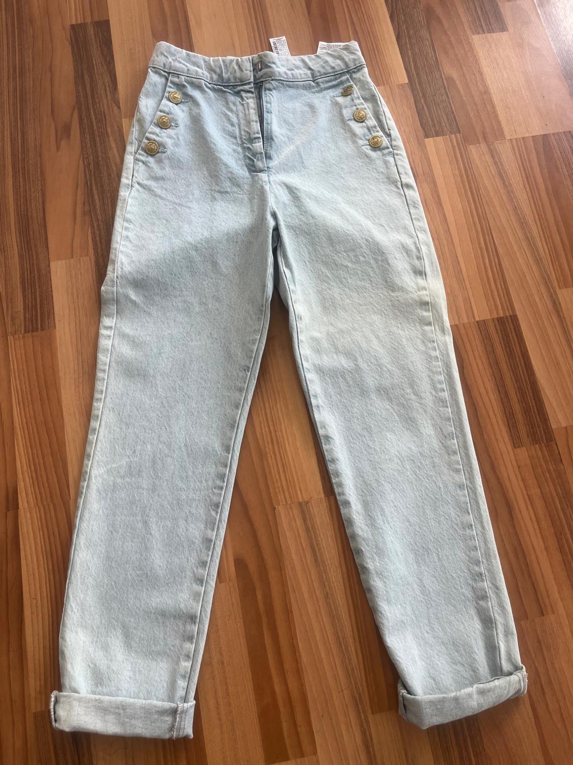 Light blue jeans with gold buttons on the sides and turn-ups, laid out on a wooden floor.