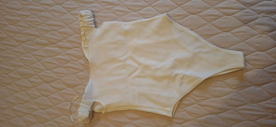 Textured white bodysuit with off-the-shoulder neckline and gathered straps, on a beige quilted bedspread.