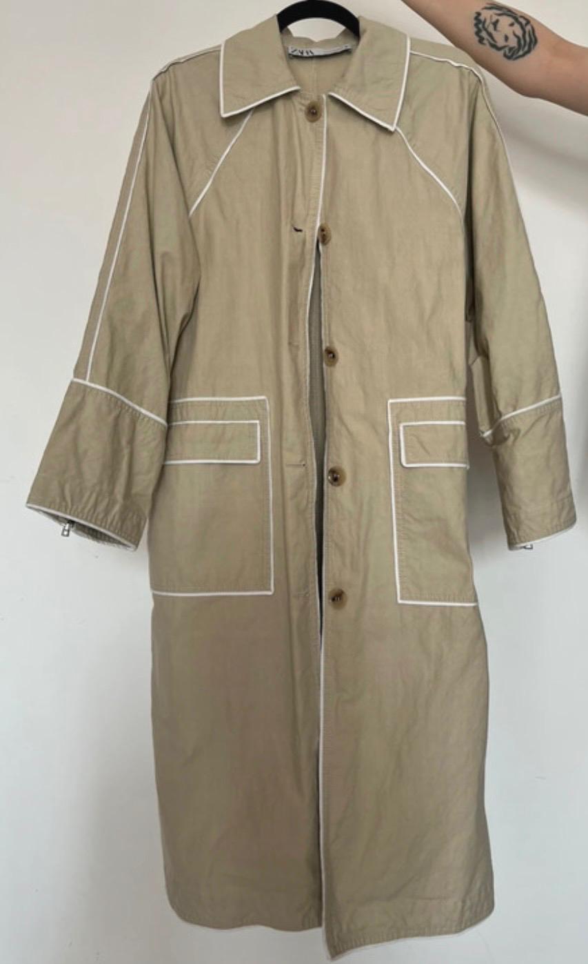 Beige coat hanging on a hanger, with visible white stitching and brown buttons. A tattooed arm is holding the hanger.