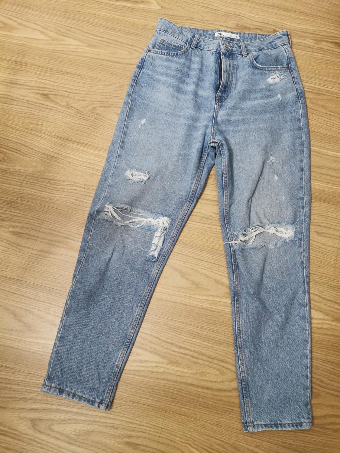Light blue denim jeans with ripped knees, laid out on a wooden floor. ZARA label on the waistband. Casual style.