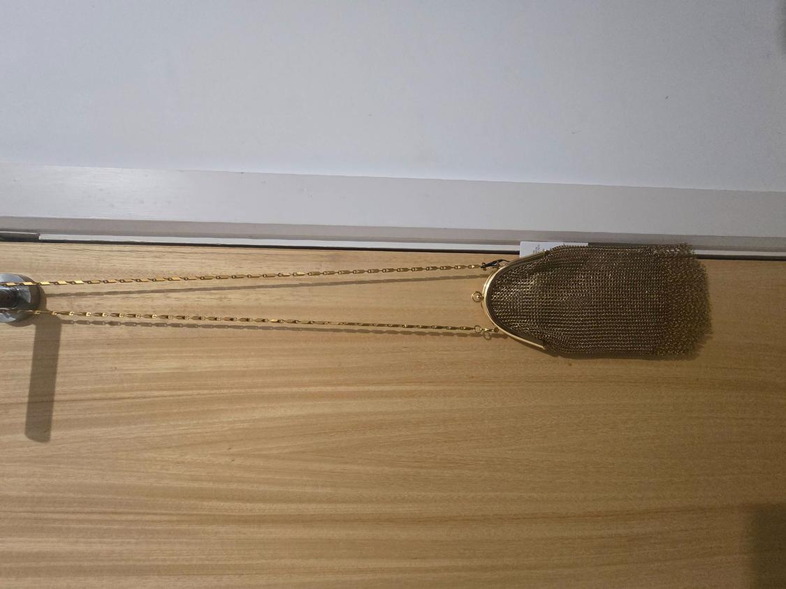 Gold mesh handbag hanging from a light wooden door. It has a long gold chain and a metal clasp.