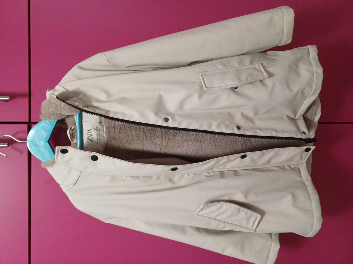 Beige jacket with brown fleece lining, hanging on a blue hanger against a pink wardrobe background. Visible label: ZARA.