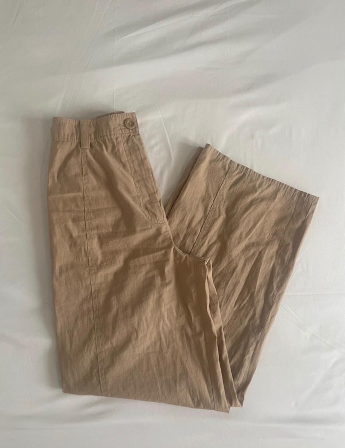 Beige wide-leg trousers with a button and front seam, folded on a crumpled white fabric. Casual style.