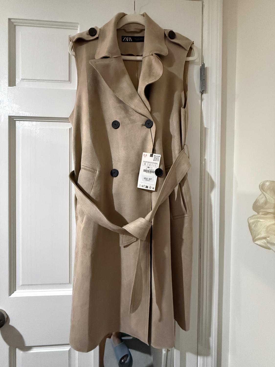 Long beige sleeveless waistcoat hanging on a white door, with black buttons and a belt. Visible tag with price and size details.