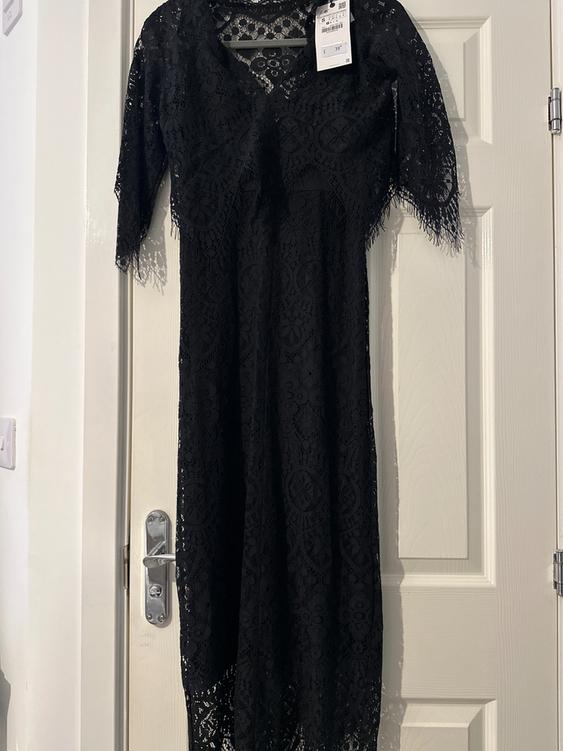 Black lace dress hanging on a white door, with three-quarter sleeves and a visible price tag on the shoulder.