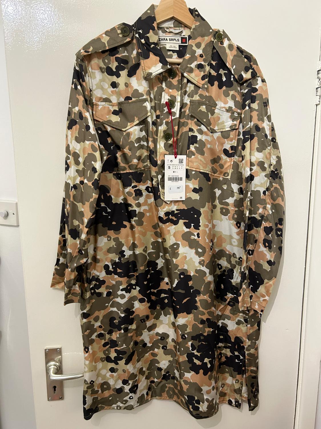 Camouflage shirt hanging on a wooden hanger. Visible label with text "ZARA SRPLS". White door background.