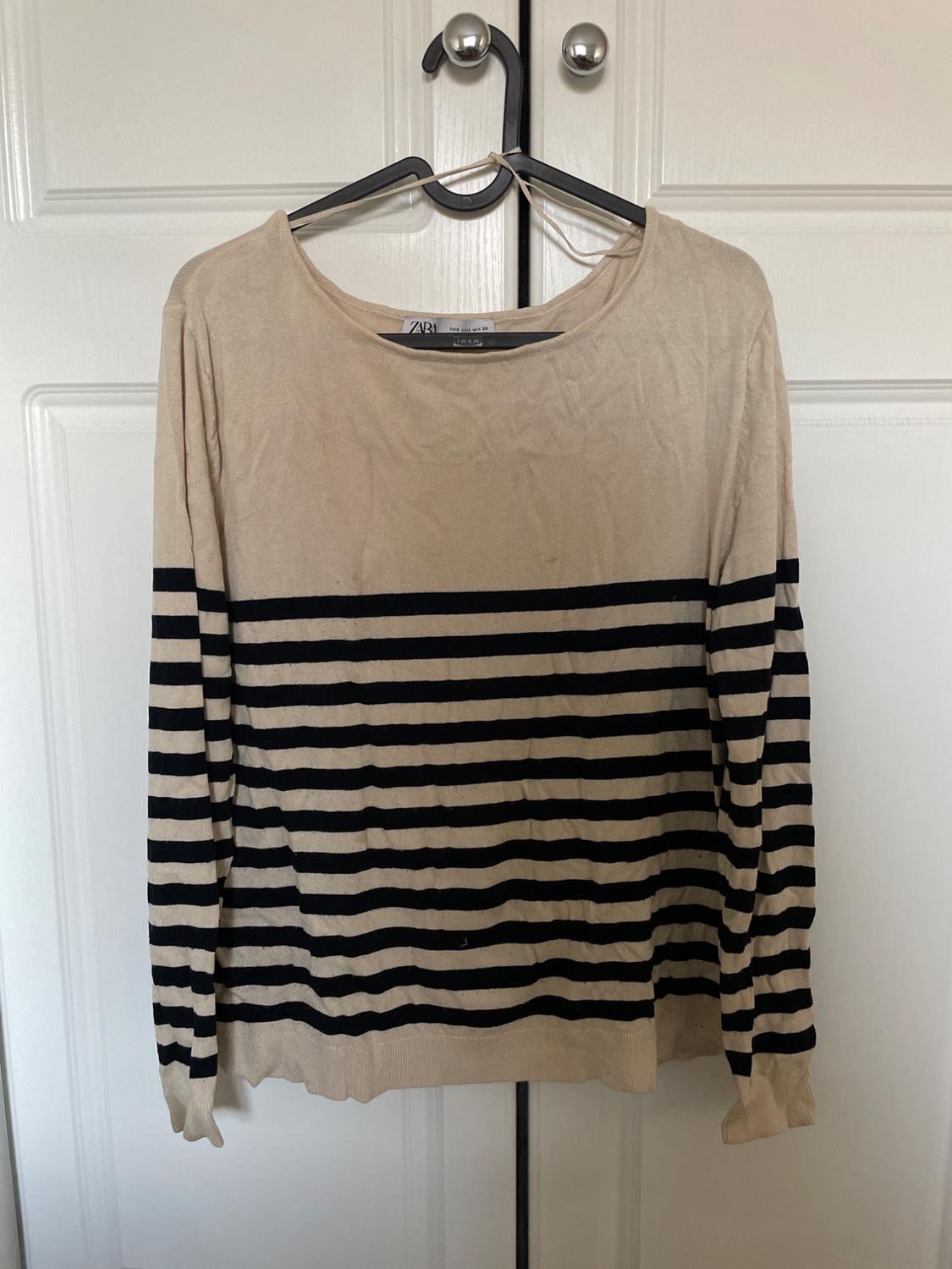 Beige long-sleeved jumper with black stripes, hanging on a hanger on a white door. ZARA label on the collar. Relaxed atmosphere.