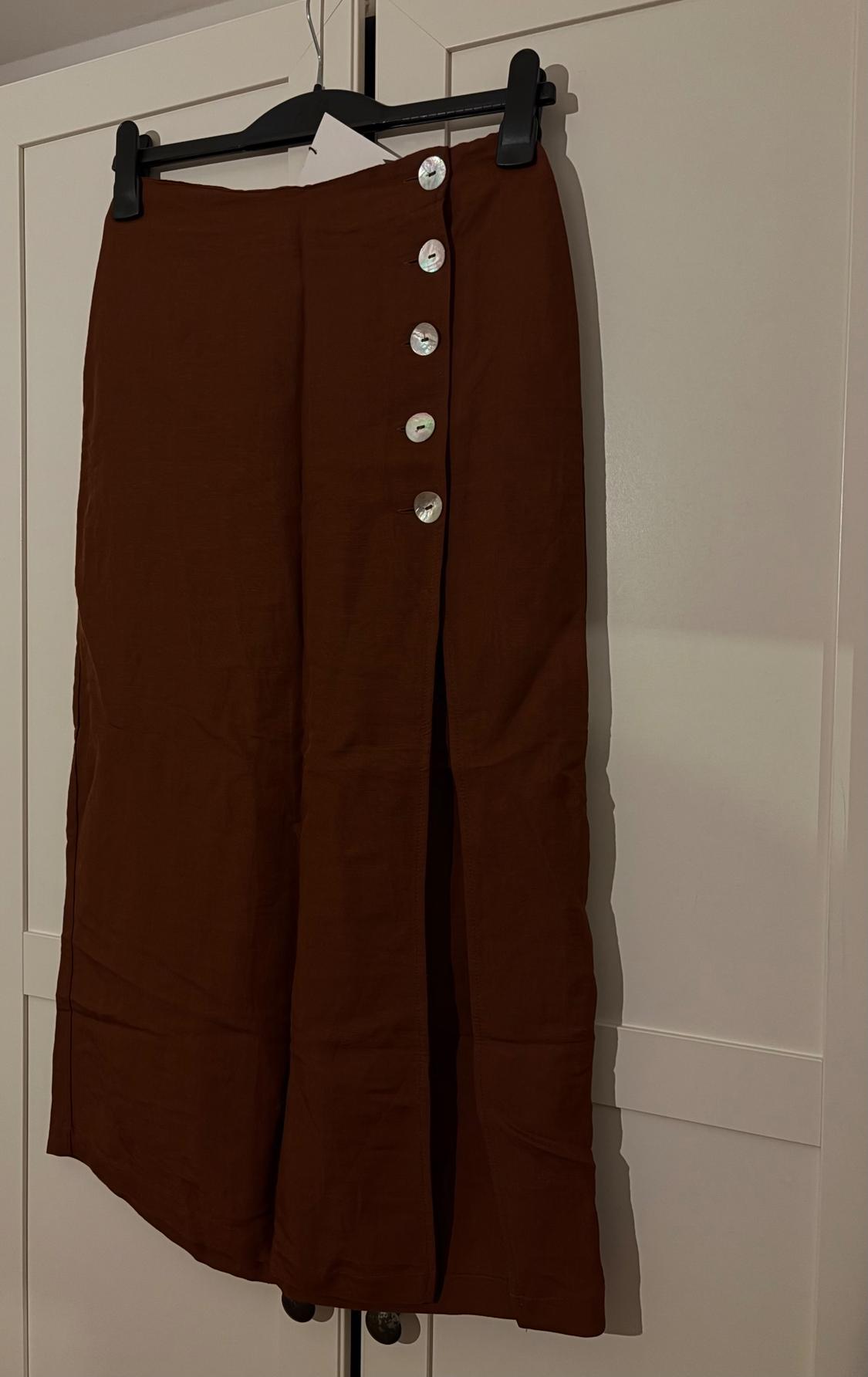 Brown skirt hanging on a black hanger, with five silver buttons at the top and a front opening. White wardrobe background.