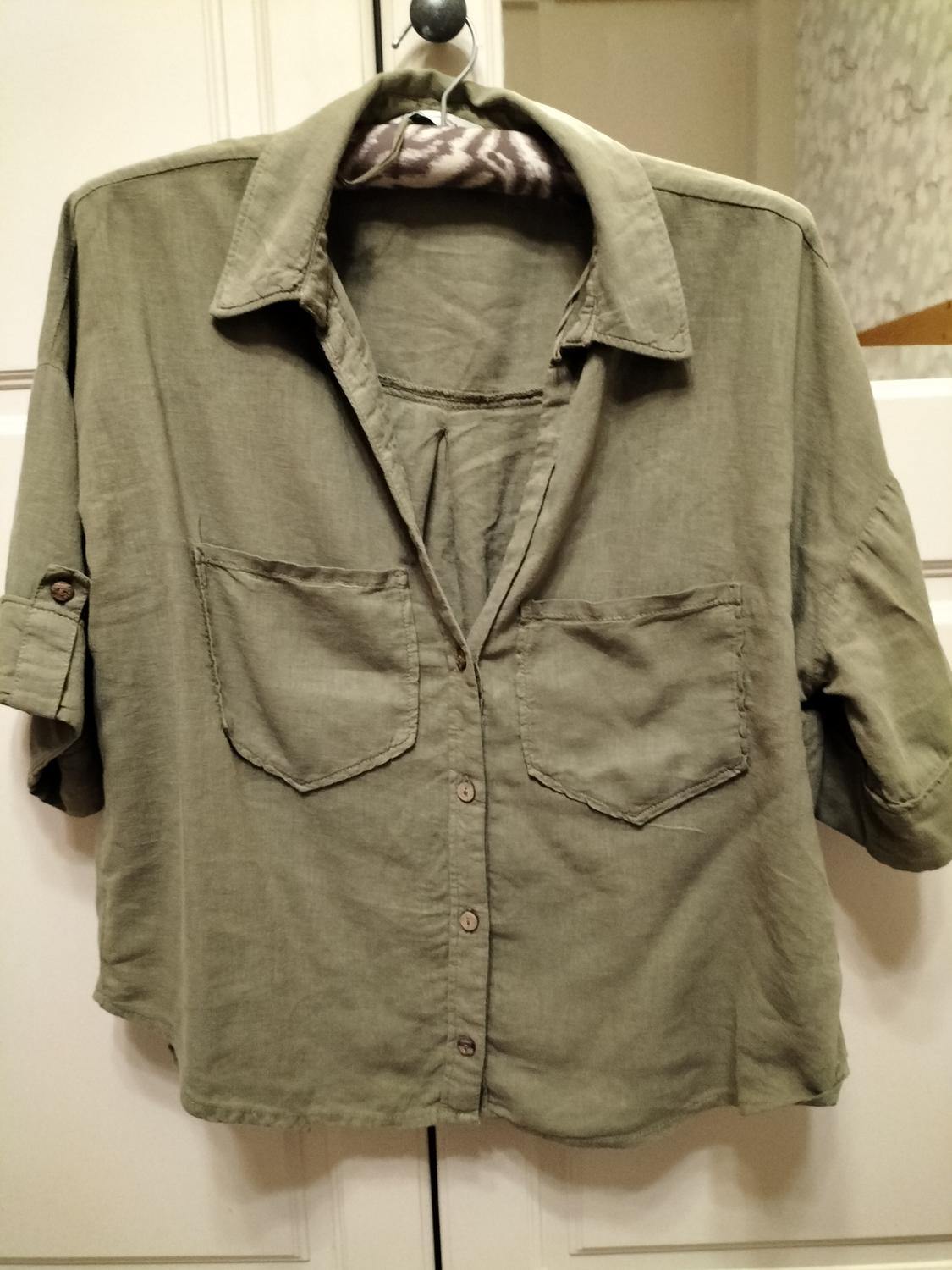 Olive green short-sleeved shirt hanging on a hanger, with two front pockets and buttons, against a white door background.
