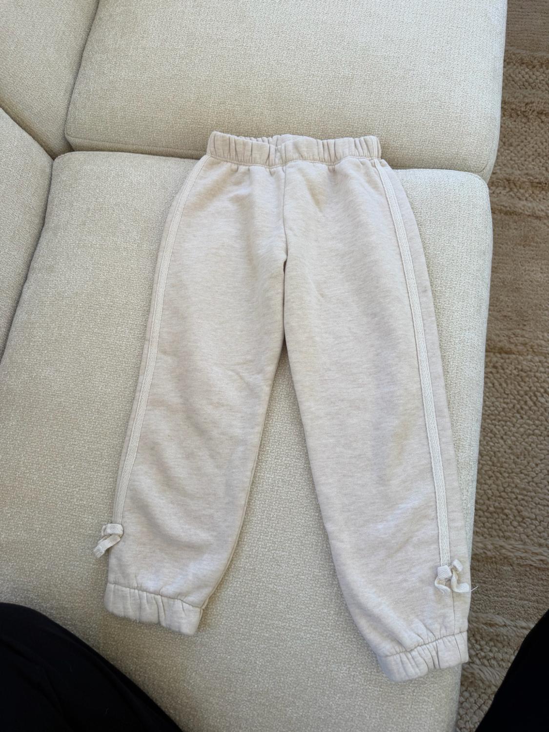 Beige track pants on a sofa. They feature an elastic waistband, cuffs at the ankles, and bow details on the sides.