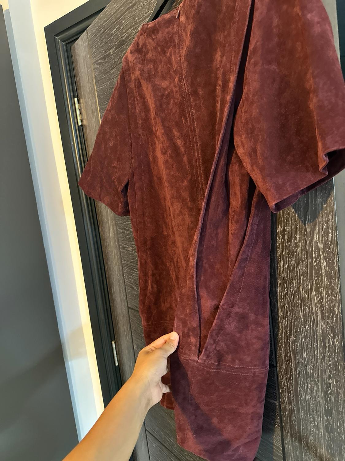 Hand showing the pocket of a maroon short-sleeved suede garment, hanging on a dark wooden door.