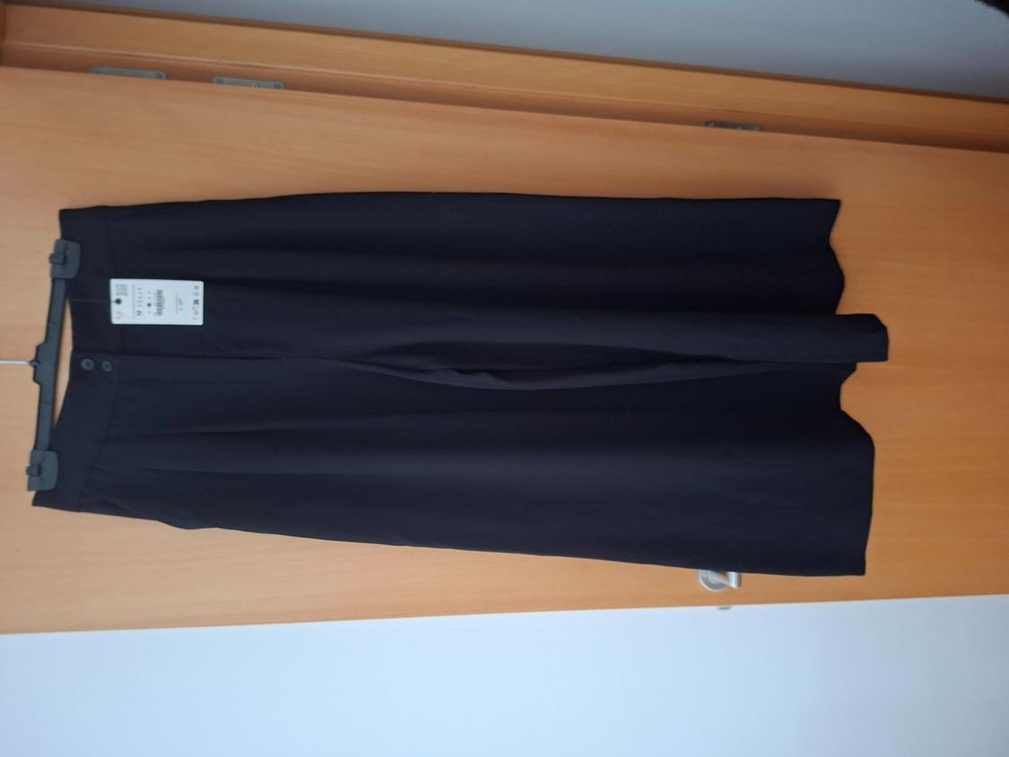 Wide black trousers hanging on a coat hanger against a wooden background. They have two buttons at the waist and a white label.
